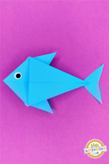Finished result of an origami fish done with blue paper and googly eyes, on top of a pink background.