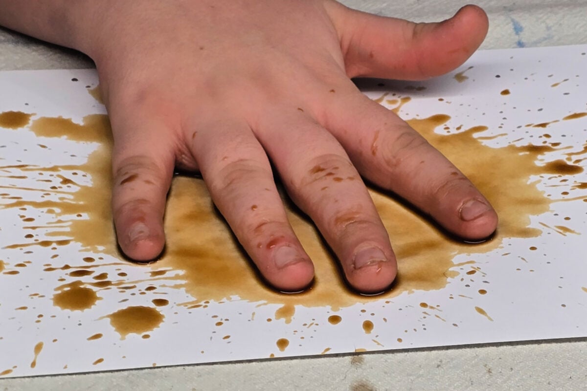 step 1 - boy patting a coffee spill with his hand to create more splatters on his paper - kids activities bog